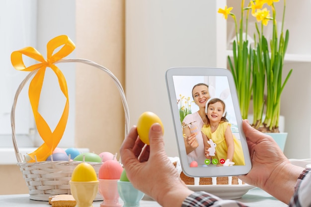 Foto eine frau hält ein digitales tablet und ein gelbes ei im arm und spricht über die osterferien per videoverbindung