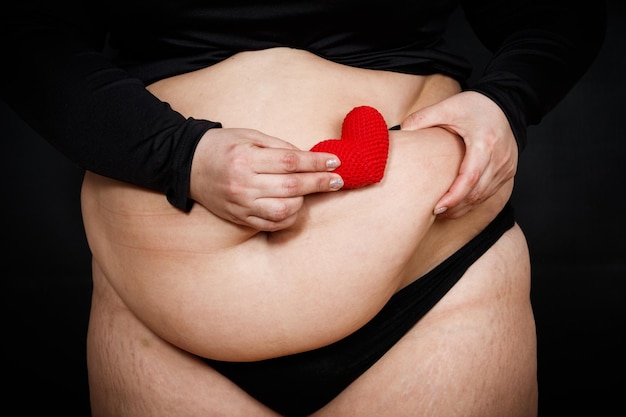 Eine Frau hält ein rotes Herz in ihren Händen auf einem dicken Bauch auf schwarzem Hintergrund fettleibige Person Körperhaltung