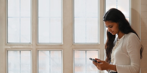 Eine Frau hält ein Smartphone in den Händen, während sie über den Fenstern des Wohnzimmers steht