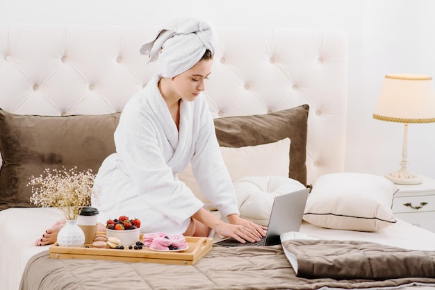 Eine Frau im Bademantel und mit Handtuch auf dem Kopf nach dem Duschen auf dem Bett mit Laptop arbeitend und lernend. Online-Arbeit zu Hause. Bleiben Sie während der Quarantäne zu Hause
