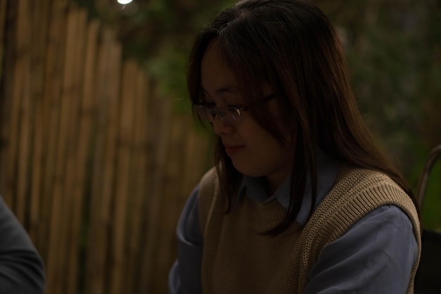 Eine Frau in blauem Hemd und Brille steht vor einem Holzzaun, an dessen Decke ein weißes Licht hängt.