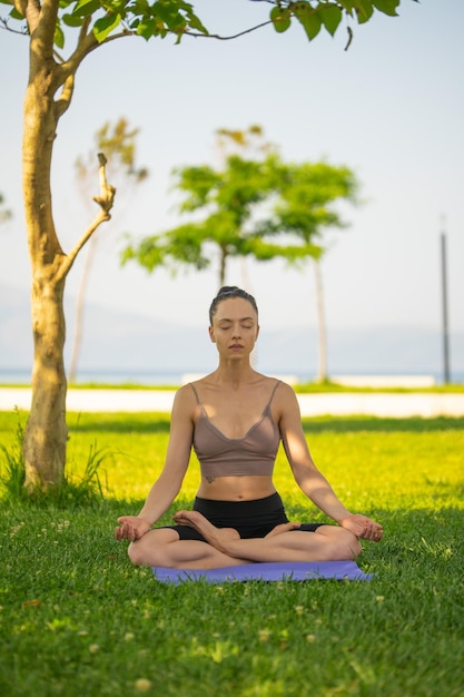 Eine Frau in braunem Sport-BH und schwarzen kurzen Hosen, die auf einer Yogamatte sitzen und Maditation mit der Hand zusammen an der Brust auf Grasboden und verschwommenem Hintergrund im Garten machen