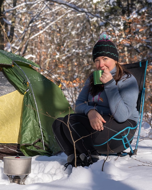 Eine Frau in der Nähe des Zeltes trinkt Tee Wickelt das Seil auf Nacht im Winterwald Liebe zur Natur Ökotourismus Sonne Wald Schnee Aktiver Lebensstil Zelt im Schnee Freude und Glück Romantik