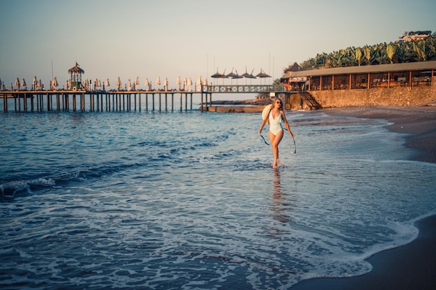 Eine Frau in einem Badeanzug und einem Hut mit Brille geht bei Sonnenuntergang am Strand entlang Das Konzept der Erholung am Meer Selektiver Fokus