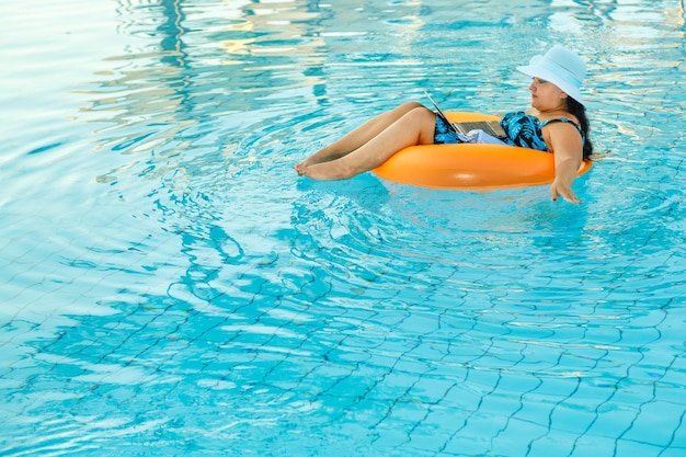 Eine Frau in einem Badeanzug und einem Sonnenhut im Pool in einem Schwimmkreis mit einem Laptop und einem Cocktail in ihren Händen.