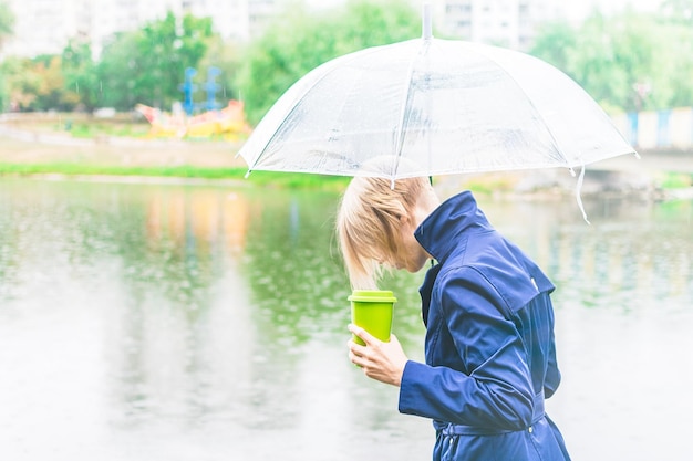 Eine Frau in einem blauen Mantel wird im Herbst in der Nähe des Sees unter einem transparenten Regenschirm mit dem Rücken gedreht