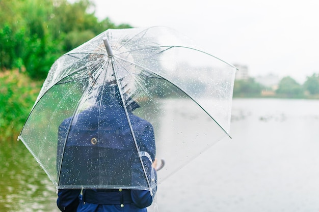 Eine Frau in einem blauen Mantel wird im Herbst in der Nähe des Sees unter einem transparenten Regenschirm mit dem Rücken gedreht