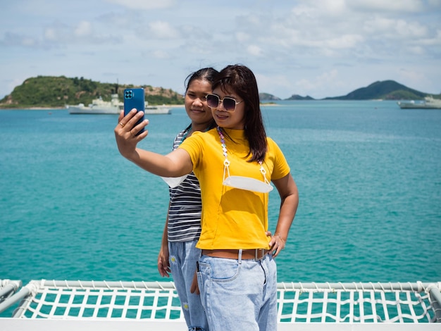 Eine Frau in einem gelben Hemd hält ein Telefon, um ein Selfie mit einem Mädchen in einem grauen Hemd mit einem Hintergrund aus blauem Wasser und kleinen Bergen zu machen Es ist Tourismus nach dem Ausbruch des Koronavirus