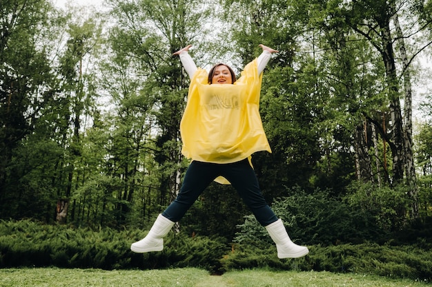 Eine Frau in einem gelben Regenmantel zeigt Emotionen und hüpft im Sommer vor dem Regen durch den Park.