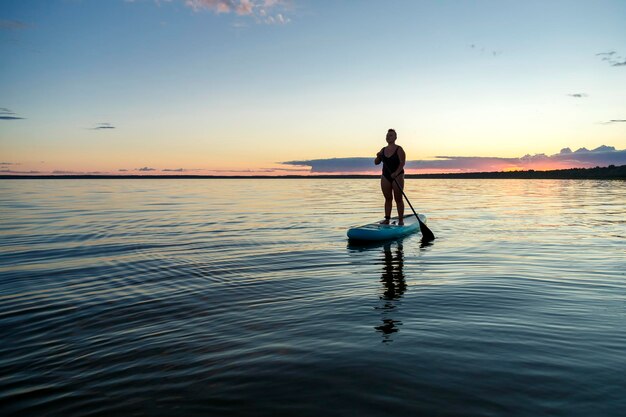 Eine Frau in einem geschlossenen Badeanzug mit Irokesenschnitt, die mit einem Ruder auf einem SUP-Board steht, schwimmt in der Abenddämmerung auf dem Wasser