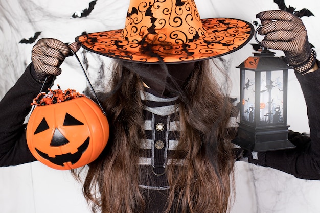 Eine Frau in einem Hexenkostüm in einer Maske hält einen Kürbis mit Süßigkeiten und eine Laterne.