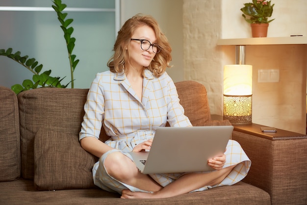 Eine Frau in einem Kleid, die mit gekreuzten Beinen auf der Couch sitzt, arbeitet aus der Ferne an einem Laptop. Ein glückliches Mädchen, das ein Webinar in ihrer Wohnung beobachtet.