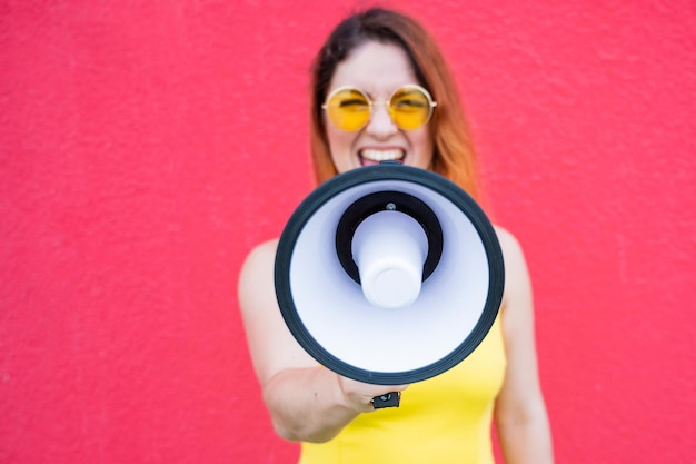 Eine Frau in einem Kleid mit Brille und Ohrringen steht im Profil auf gelbem Hintergrund und schreit in ein Megaphon. Porträt eines Mädchens mit einem Lautsprecher. Eine Dame schreit ein Gerät zur Tonverstärkung an
