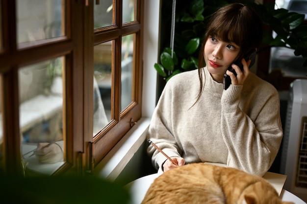 Eine Frau in einem kuscheligen Pullover telefoniert mit ihrer Katze und sitzt an einem Tisch am Fenster