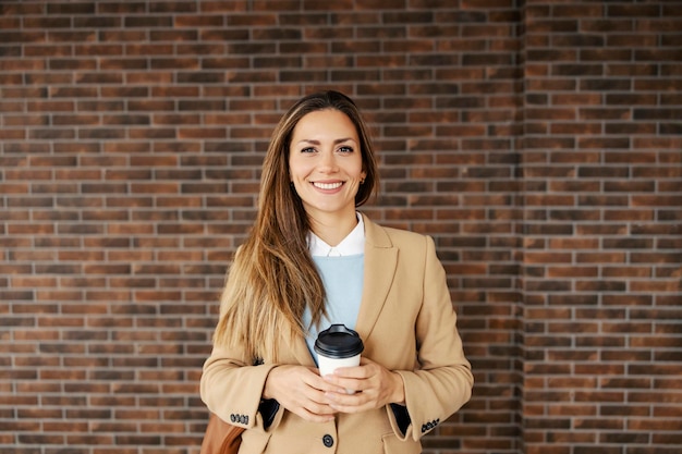 Eine Frau in einem Mantel, die auf der Straße steht und Kaffee zum Mitnehmen hält