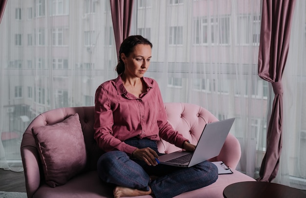 Eine Frau in einem rosa Hemd sitzt mit einem Laptop auf dem Sofa und arbeitet