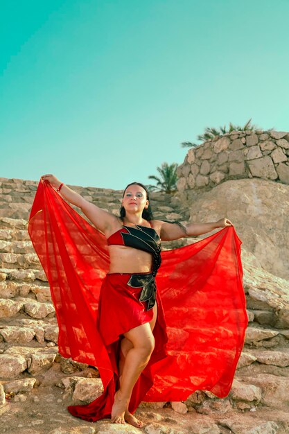 Eine Frau in einem roten Anzug tanzt einen orientalischen Tanz auf einer Steintreppe gegen den Himmel. vertikales Foto