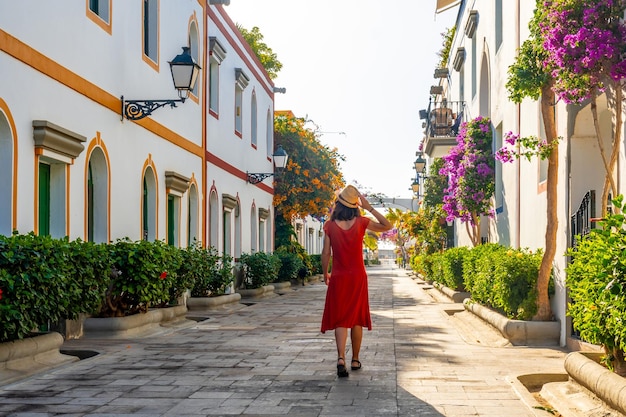 Eine Frau in einem roten Kleid geht mit einem Hut eine schmale Straße entlang