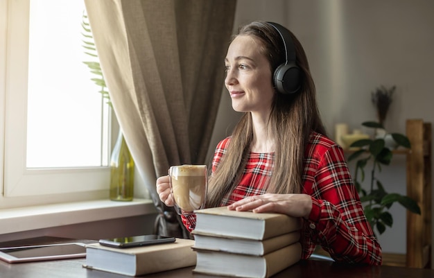 Eine Frau in einem roten Kleid hört Hörbuch mit Kopfhörern und trinkt Kaffee am Fenster Konzept der modernen technologischen Bildung und des Studiums von Büchern mit Freude