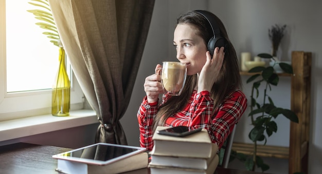 Eine Frau in einem roten Kleid hört Hörbuch mit Kopfhörern und trinkt Kaffee Das Konzept der modernen technologischen Bildung und das Studium von Büchern mit Freude