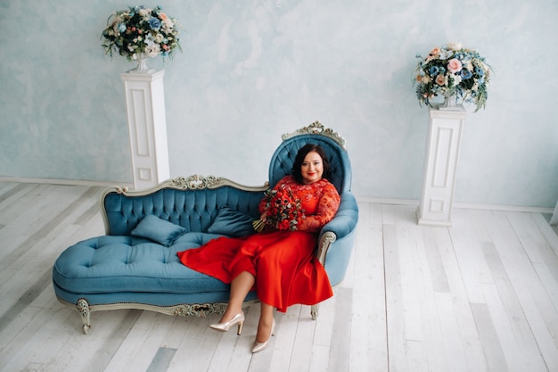 Eine Frau in einem roten Kleid sitzt auf einem Sofa und hält im Innenraum einen Strauß roter Rosen und Erdbeeren.