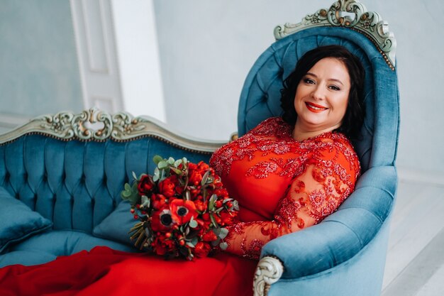 Eine Frau in einem roten Kleid sitzt auf einem Sofa und hält im Inneren einen Strauß roter Rosen und Erdbeeren.