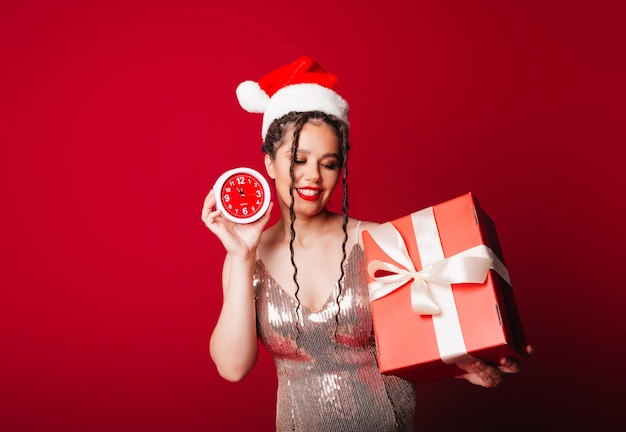 Eine Frau in einem roten Weihnachtshut mit Dreadlocks hält ein Geschenk auf rotem Hintergrund Isolieren Sie eine Frau in einem eleganten Kleid Neujahr Weihnachten