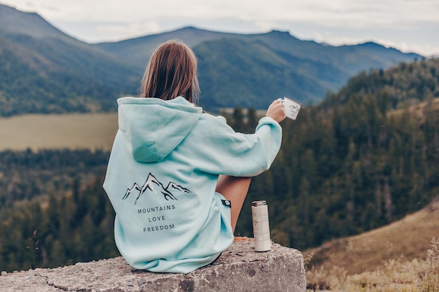 Eine Frau in einem schönen blauen Oversize-Hoodie mit Aufdruck sitzt auf einem grauen Felsen und blickt auf eine Alm und trinkt heißen Tee aus einem Becher in ihren Händen. Bewundern Sie die natürliche Umgebung.