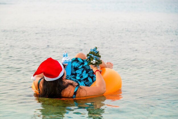 Eine Frau in einem Schwimmring in einer Weihnachtsmannmütze mit einem Cocktail und einem kleinen Weihnachtsbaum