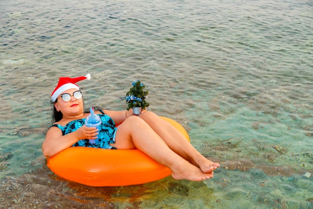 Eine Frau in einem Schwimmring in einer Weihnachtsmannmütze mit einem Cocktail und einem Weihnachtsbaum in der Hand