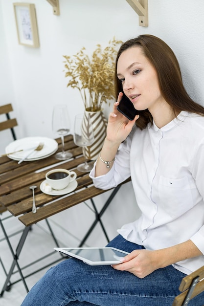 Eine Frau in einem weißen Hemd und Jeans sitzt in einem Café mit einem Tablet in ihren Händen Weiblicher Büroangestellter in der Mittagspause am Telefon sprechen Manager bei einem Geschäftstreffen in einem Restaurant trinkt Kaffee