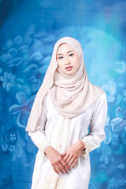Eine Frau in einem weißen Hijab steht vor einem blauen Hintergrund mit Blumen.