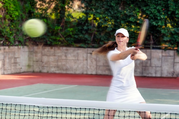 Eine Frau in einem weißen Kleid und einer Baseballmütze spielt im Sommer Rasentennis auf einem Tennisplatz, schlägt den Ball mit einem Schläger oder macht einen Platz, Sport und Freizeit.