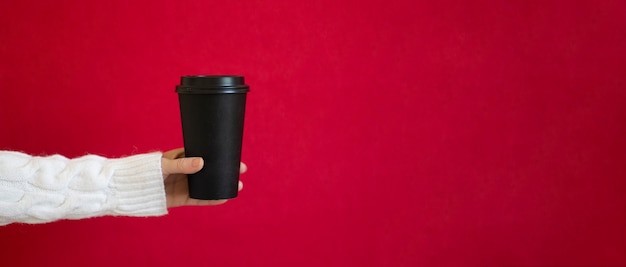 Eine Frau in einem weißen Pullover hält einen schwarzen Pappbecher für Kaffee auf rotem Grund.