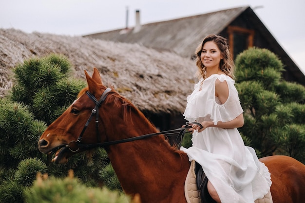 Eine Frau in einem weißen Sommerkleid reitet auf einem Pferd in der Nähe eines Bauernhofs