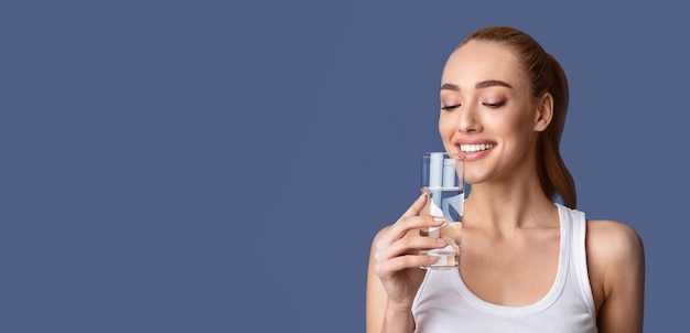 Eine Frau in einem weißen Tanktop trinkt Wasser aus einem Glas, während sie mit ihren Augen auf das Wasser schaut