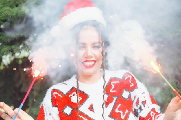 Eine Frau in einem Winterpullover mit Weihnachtsmannmütze und roten Lippen hält eine Wunderkerze vor dem Hintergrund von Weihnachtsbäumen Neujahr Weihnachten