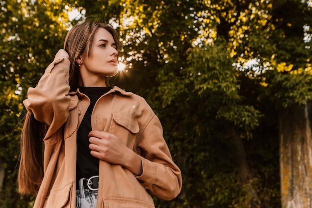 Eine Frau in einer braunen Jacke steht vor einem Baum und schaut in die Kamera