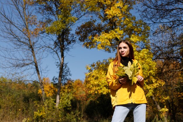 Eine Frau in einer gelben Jacke mit Herbstlaub auf einem hellen Herbsthintergrund bei einem Spaziergang im Park