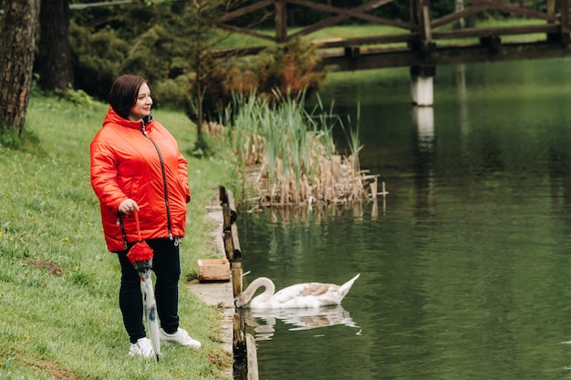 Eine Frau in einer roten Jacke und mit einem Regenschirm geht durch einen Sommerpark in der Nähe eines Sees mit Schwänen.