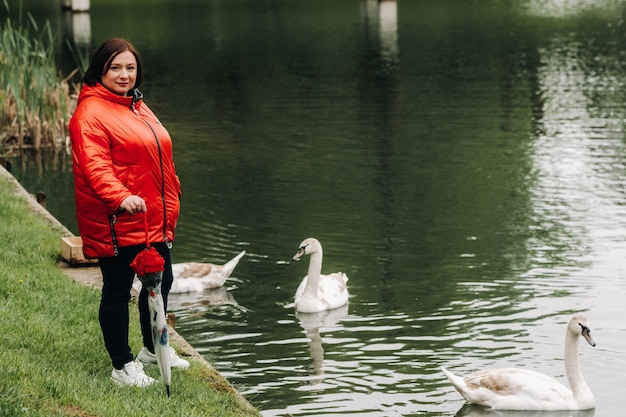 Eine Frau in einer roten Jacke und mit einem Regenschirm geht durch einen Sommerpark in der Nähe eines Sees mit Schwänen.