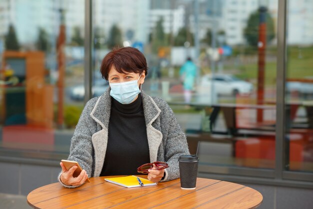 Eine Frau in einer Schutzmaske sitzt an einem Tisch in einem Sommercafé und hält ein Smartphone in der Hand