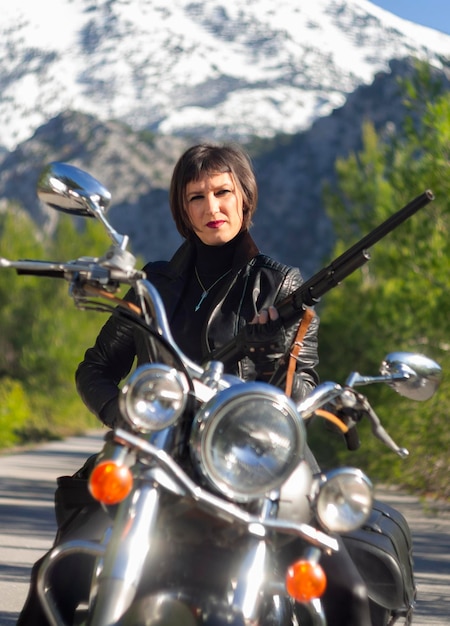 Eine Frau in einer schwarzen Bikerjacke aus Leder mit einem Karabinergewehr auf einem Chopper-Motorrad in Griechenland