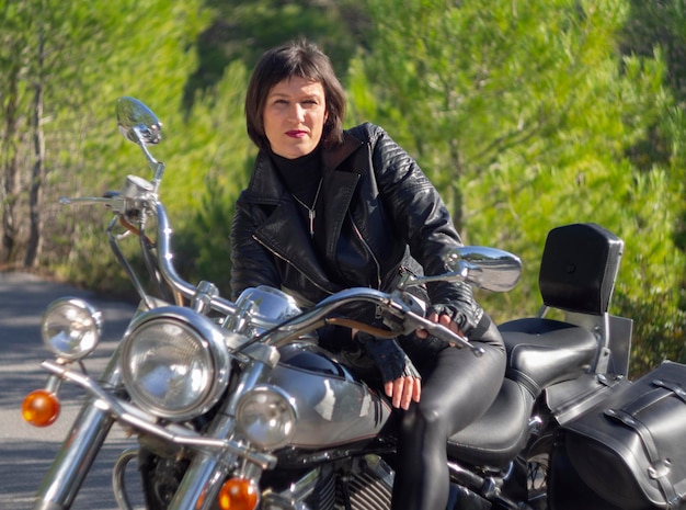 Eine Frau in einer schwarzen Leder-Bikerjacke auf einem Chopper-Motorrad in Griechenland auf einer Straße im Wald
