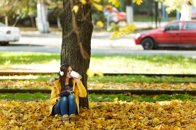 Eine Frau in gelbem Mantel und Jeans sitzt mit einer Tasse Kaffee oder Tee und hört Musik unter einem Baum mit einem Tablet in den Händen und Kopfhörern im Herbststadtpark an einem warmen Tag. Goldene Herbstblätter.