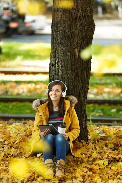 Eine Frau in gelbem Mantel und Jeans sitzt mit einer Tasse Kaffee oder Tee und hört Musik unter einem Baum mit einem Tablet in den Händen und Kopfhörern im Herbststadtpark an einem warmen Tag. Goldene Herbstblätter.