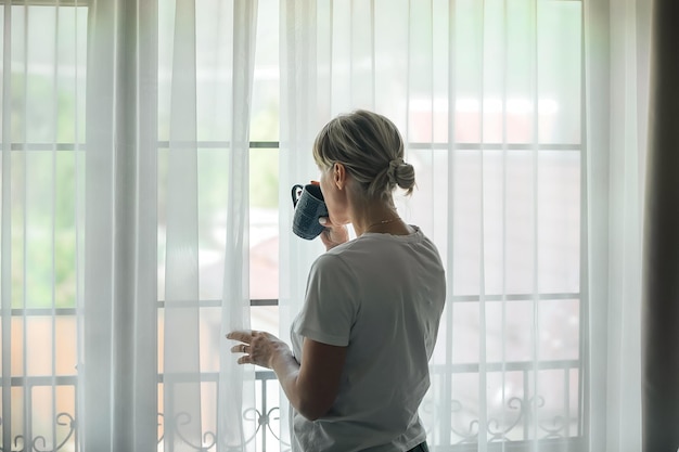 eine frau in jeans und t-shirt trinkt zu hause am fenster kaffee und schaut hinein