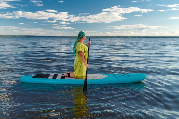 Eine Frau in Kleid und Schal auf einem Brettchen schwimmt kniend auf dem See