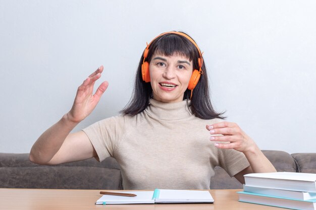 Eine Frau in orangefarbenen Kopfhörern, die an einer Online-Diskussion teilnimmt und aktiv gestikuliert, während sie an einem Tisch mit Notizbüchern und Büchern sitzt