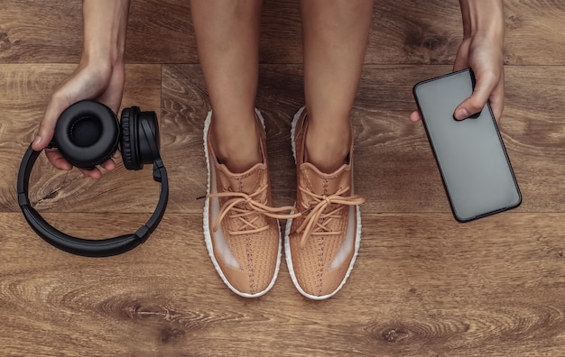 Eine Frau in Sportschuhen hält ein Smartphone und kabellose Kopfhörer in den Händen, während sie auf dem Boden sitzt. Gesundes Lebensstilkonzept, Training. Ansicht von oben